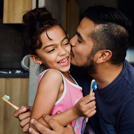 Man kissing his daughter on the cheek as they both hold toothbrushes